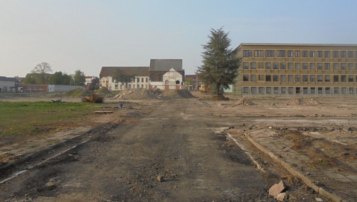 Een vlak, leeg terrein na een afbraak, met de kerk en huizen van Eeklo op de achtergrond